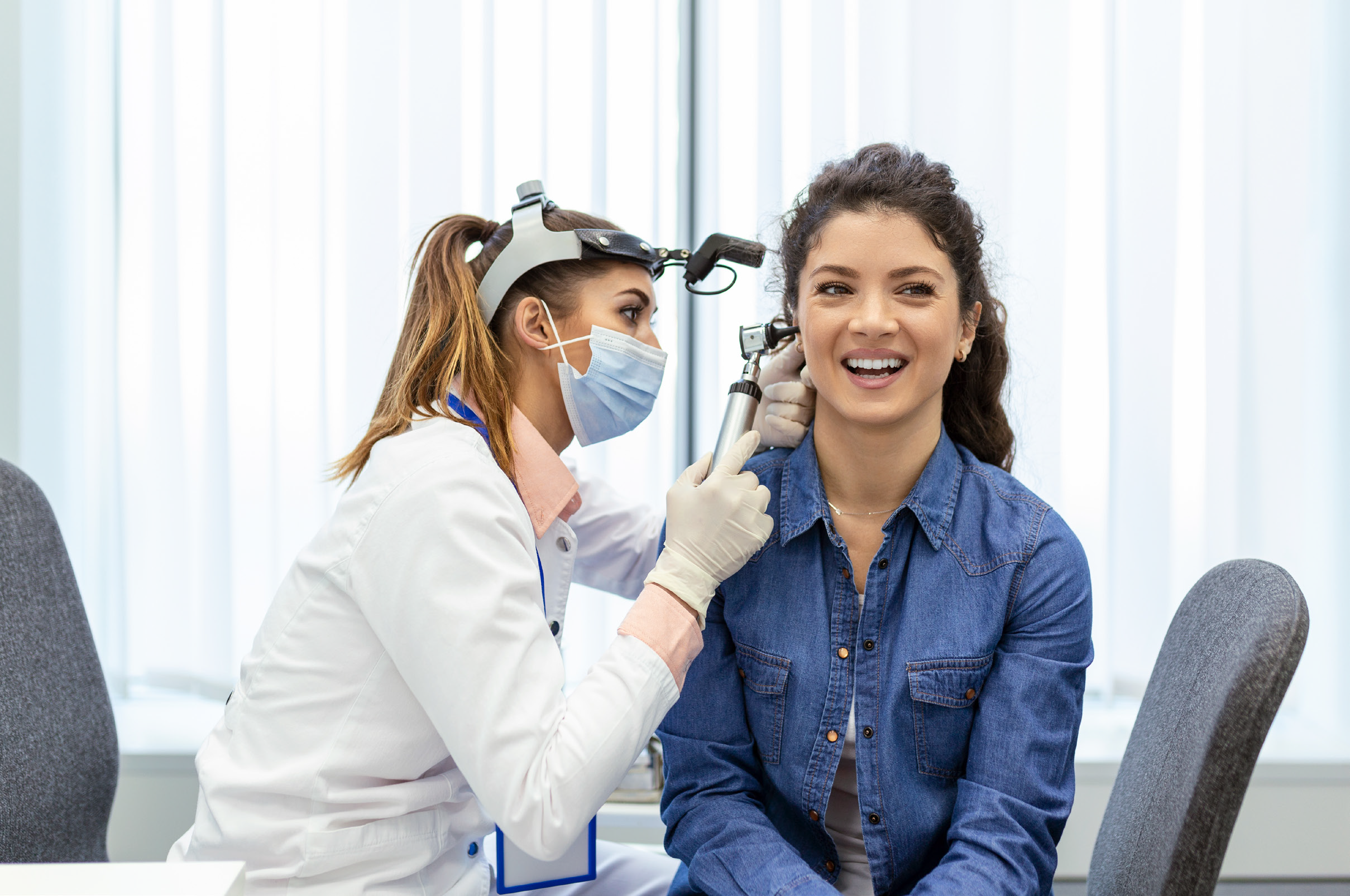 Female audiologist in a white coat and blue surgical mask performing a hearing examination on a smiling patient wearing a denim shirt. The audiologist is using an otoscope to examine the patient's ear in a bright, modern medical office with large windows in the background.